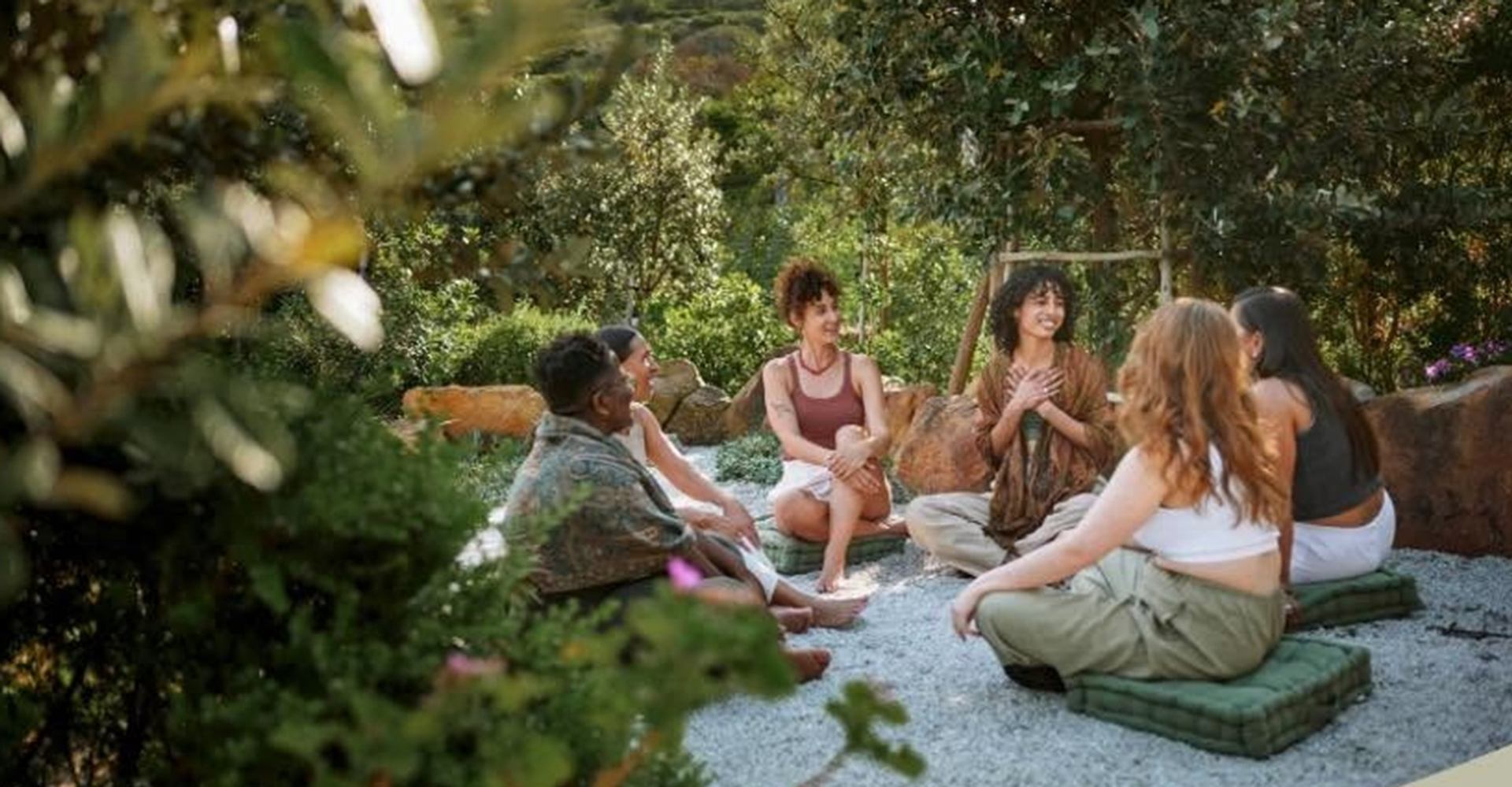 Group of people sitting on cushions in a garden, talking in a circle.