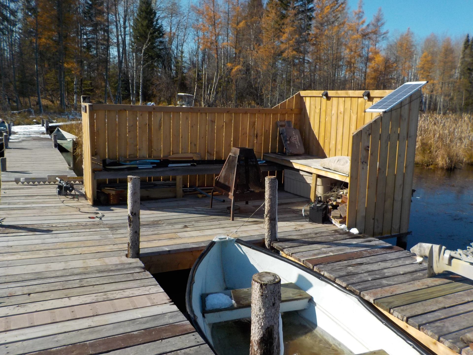 Dock with seating and solar panel beside a boat on a calm lake with autumn trees.