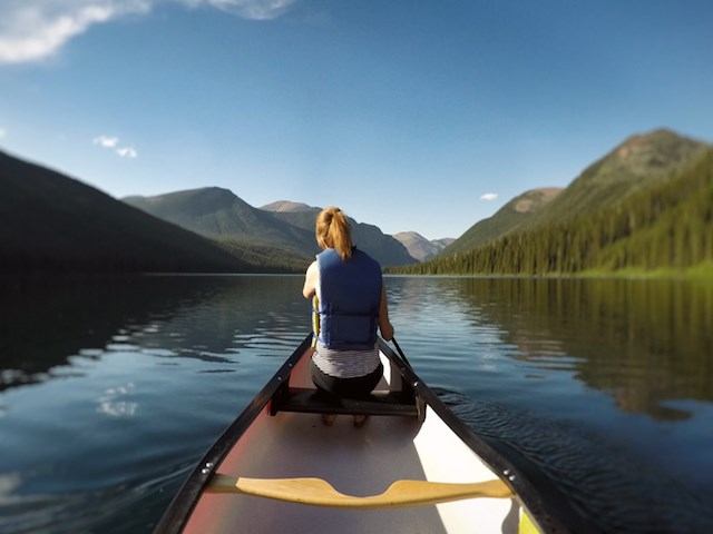 Person canoeing on calm lake with mountain reflections and clear skies.