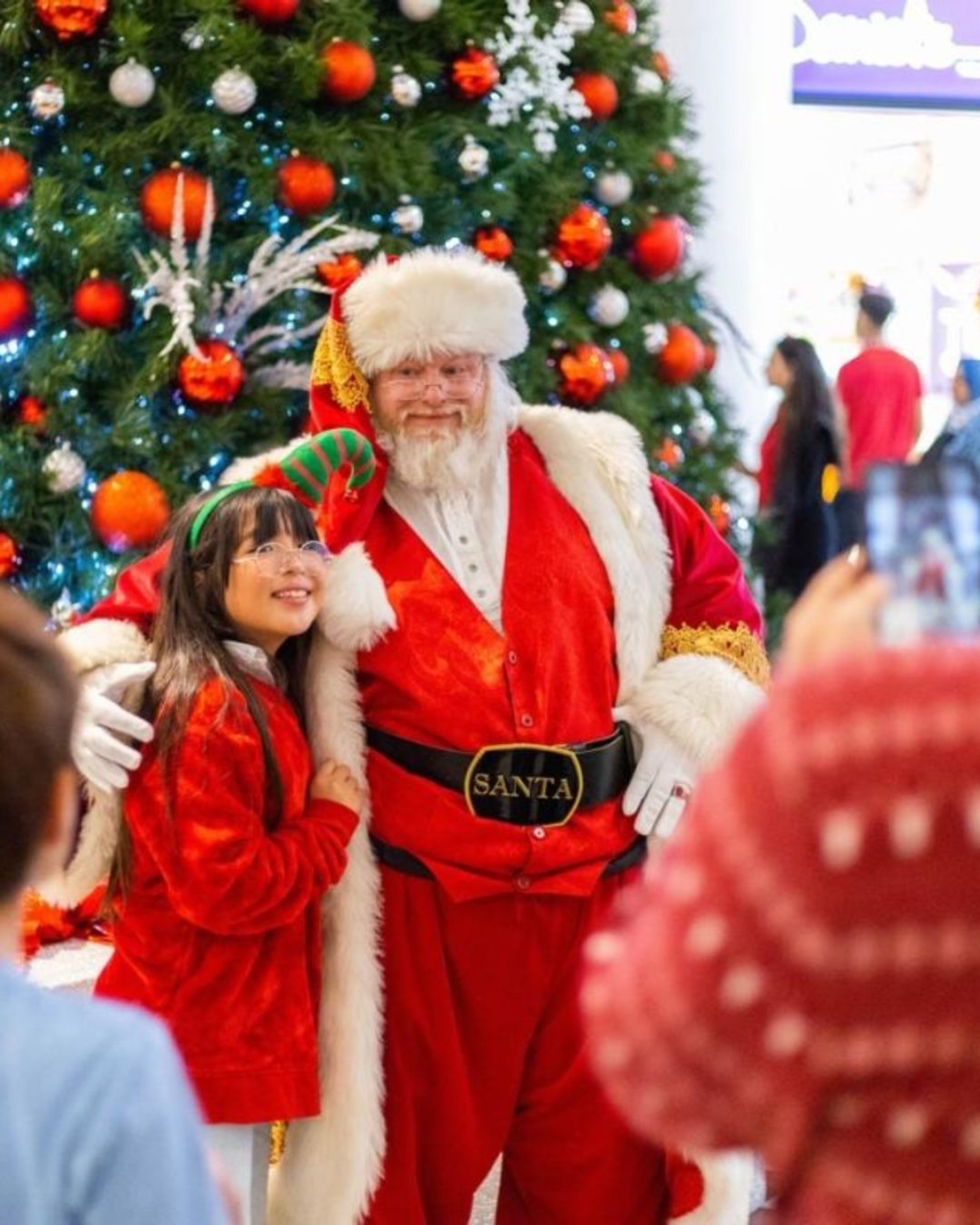 Santa poses with a person in an elf hat beside a decorated Christmas tree.