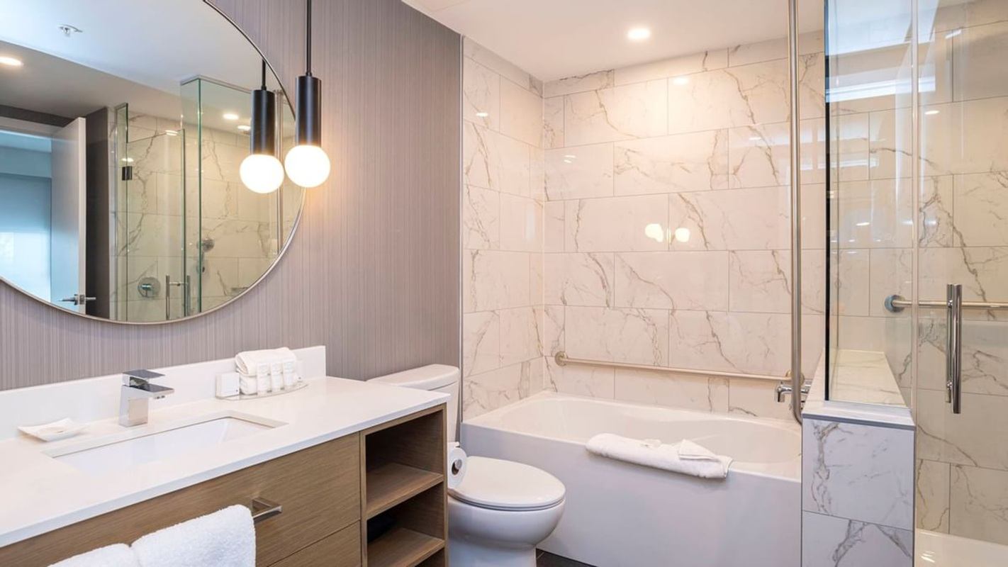 Contemporary hotel bathroom with marble tile walls, bathtub with glass shower panel, modern vanity with sink, round mirror, and pendant lighting.
