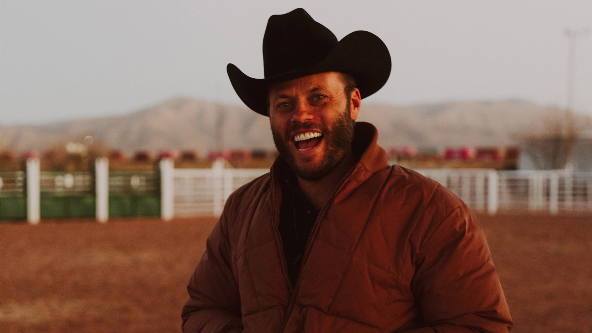 Person in cowboy hat and jacket standing in an outdoor arena.