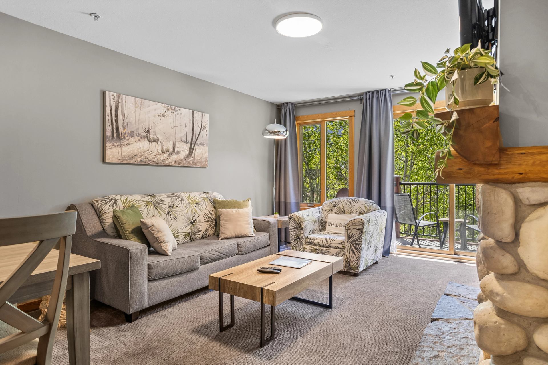 Living room with sofa, armchair, coffee table, wall art, and large windows overlooking greenery.