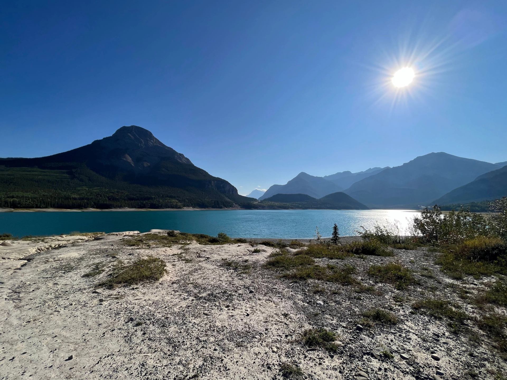 Rocky shoreline with turquoise lake, forested mountains and bright sun in clear sky.