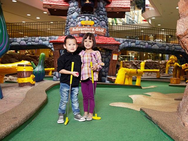 Children hold mini-golf clubs on indoor course beneath Adventure Golf sign.