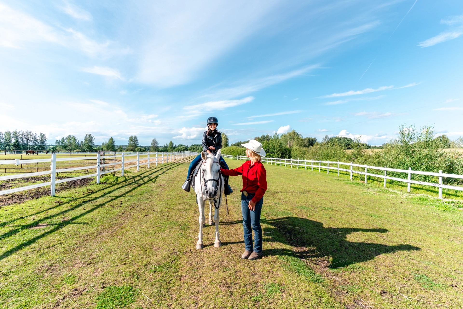 Horse rider on white horse with instructor in red shirt at fenced grassy paddock.