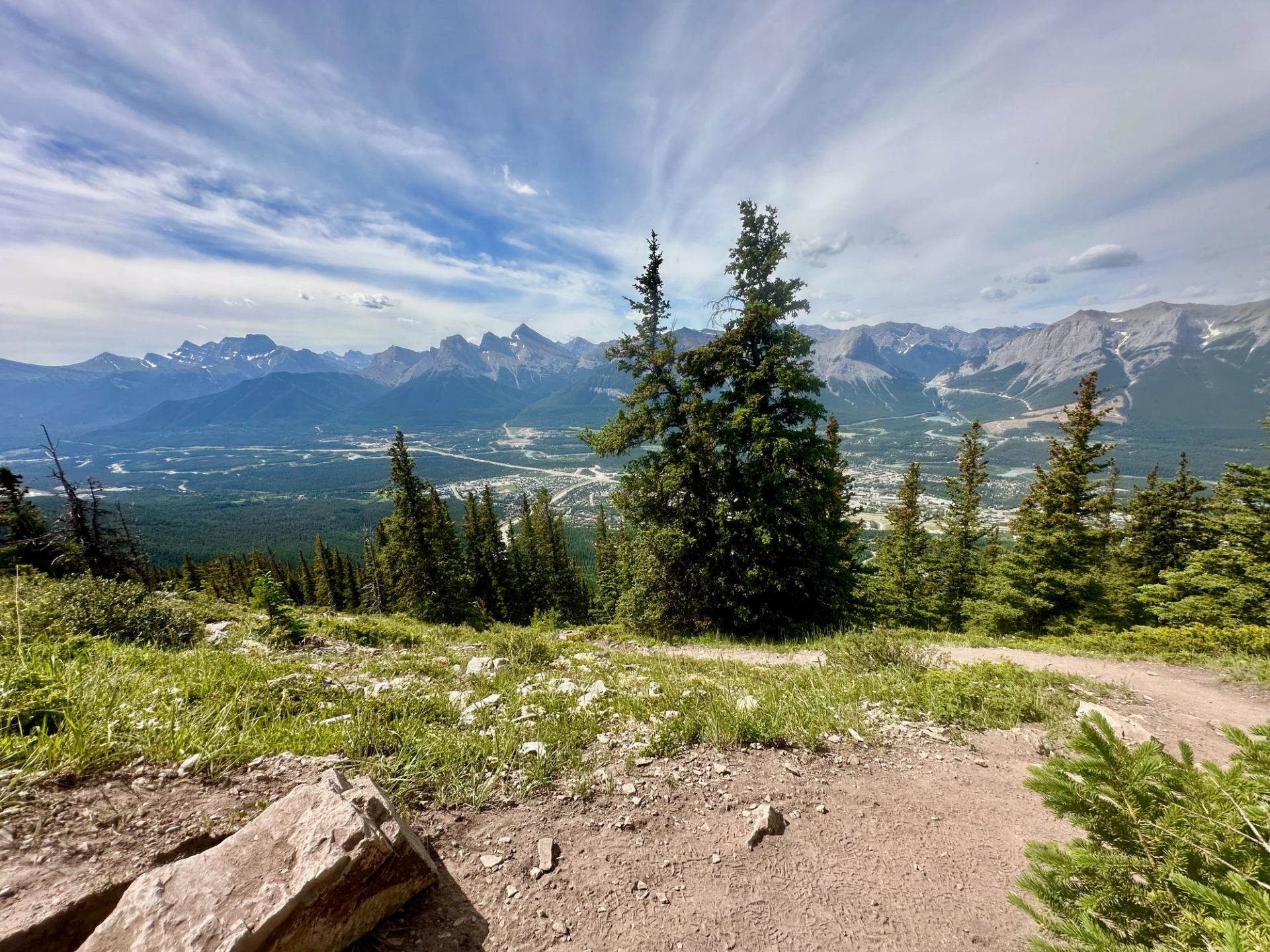 Mountain trail overlooking forested valley, distant town and rugged peaks under streaked clouds.