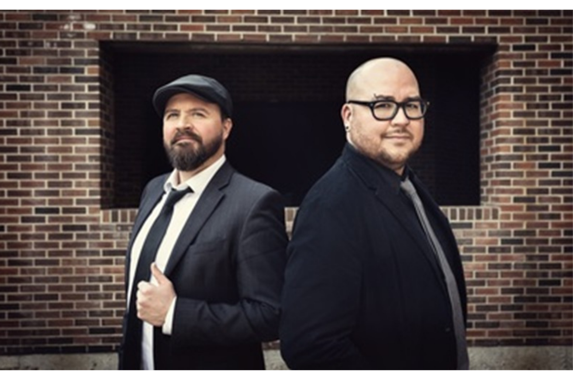 Two people in dark suits stand back-to-back before a brick wall.