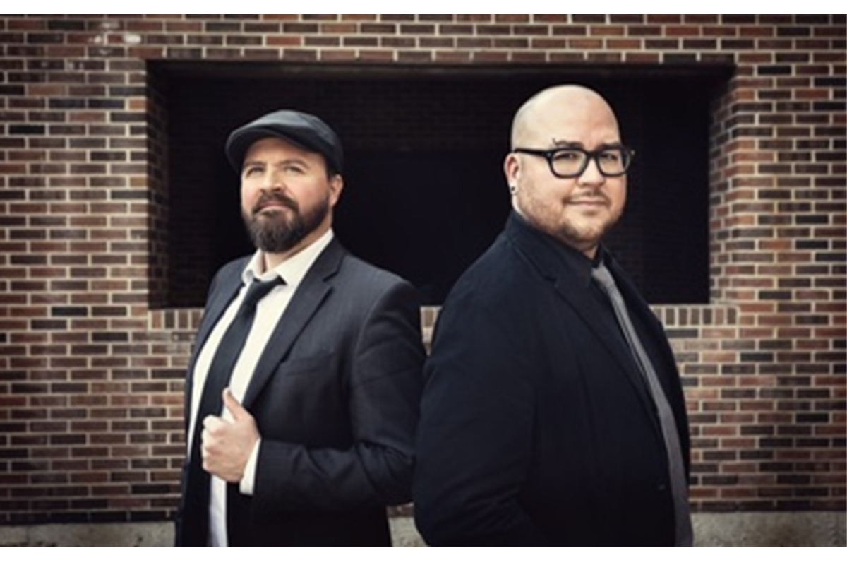 Two people in dark suits stand back-to-back before a brick wall.
