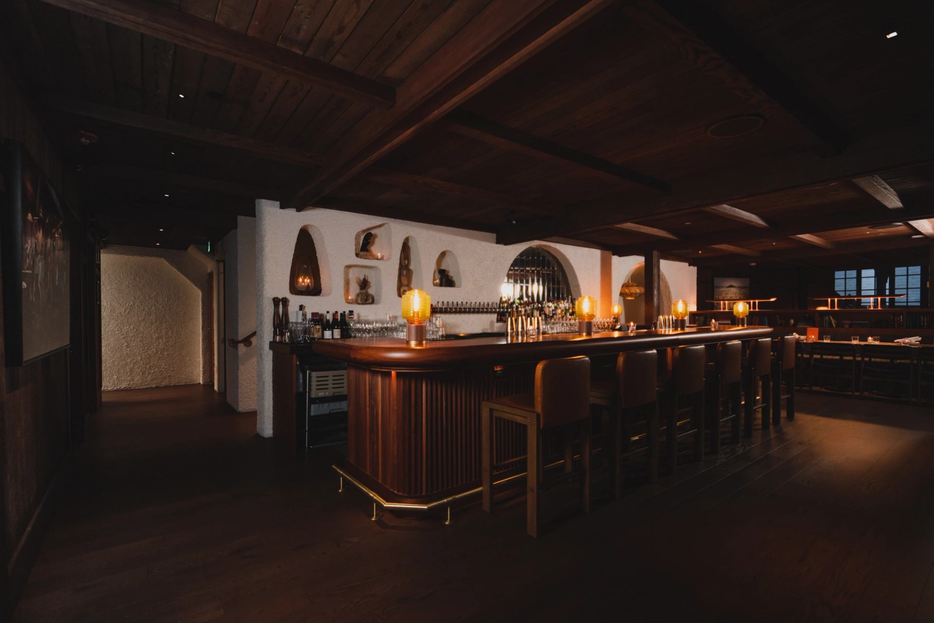 A dimly lit, elegant bar interior with a long wooden counter, bar stools, and warm decorative lighting.