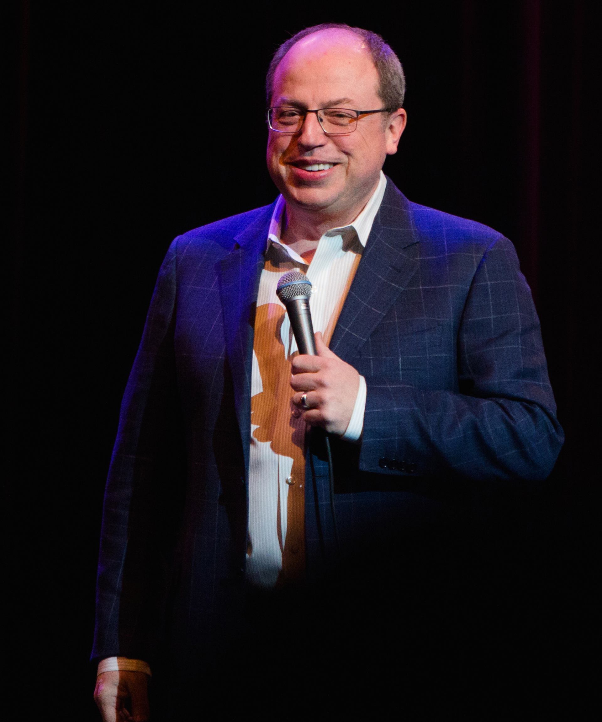 Comedian in suit jacket holds microphone on dark stage under bright spotlight.
