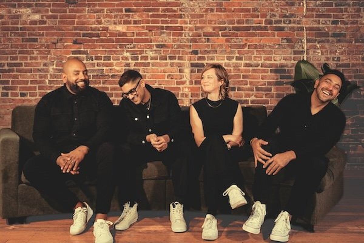 Musicians sit on stage in front of brick wall, smiling, with instruments nearby.
