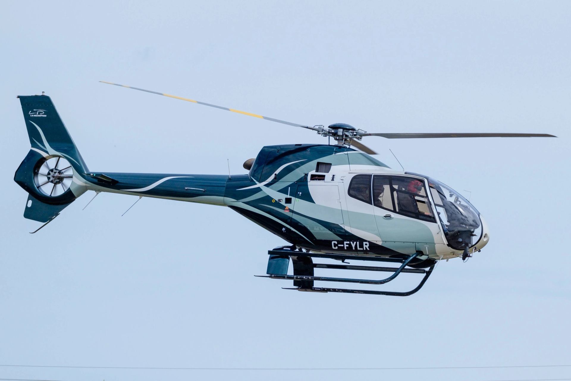 Helicopter with teal and white livery flying in clear blue sky, registration C-FYLR visible.