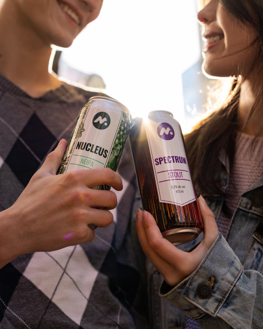 Two people clinking Meta Brewery cans labeled Nucleus and Spectrum in sunlight.