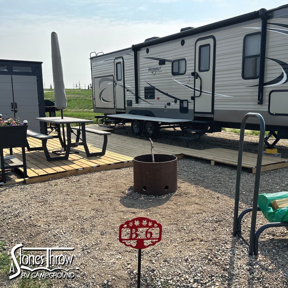 A parked RV on gravel next to a wooden deck with patio furniture, closed umbrella, and fire pit for coziness.