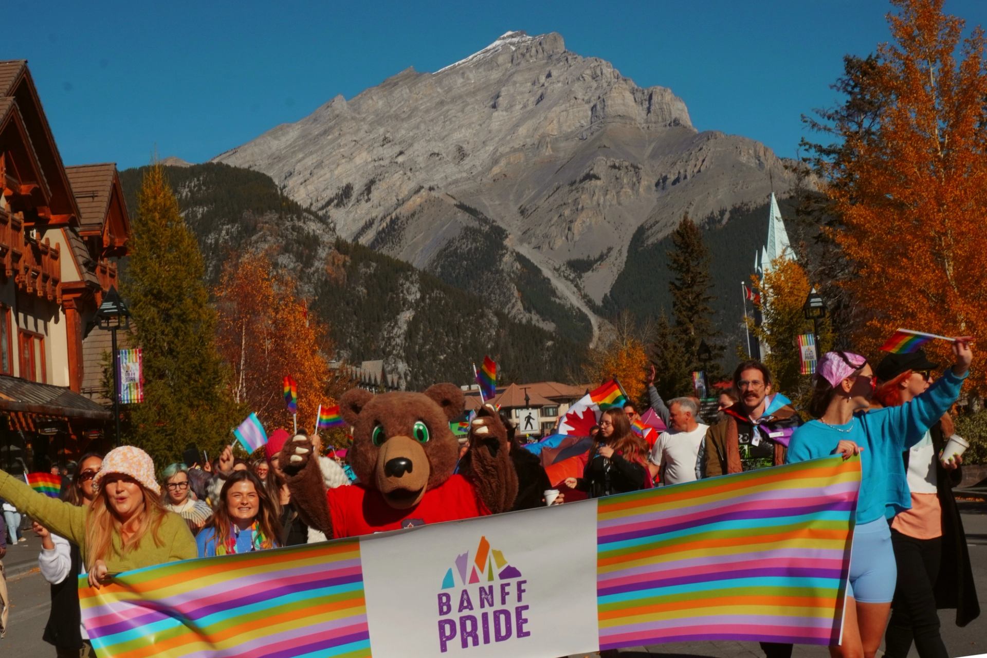 Banff Pride marchers carry a rainbow banner beneath a mountain and autumn-coloured trees.