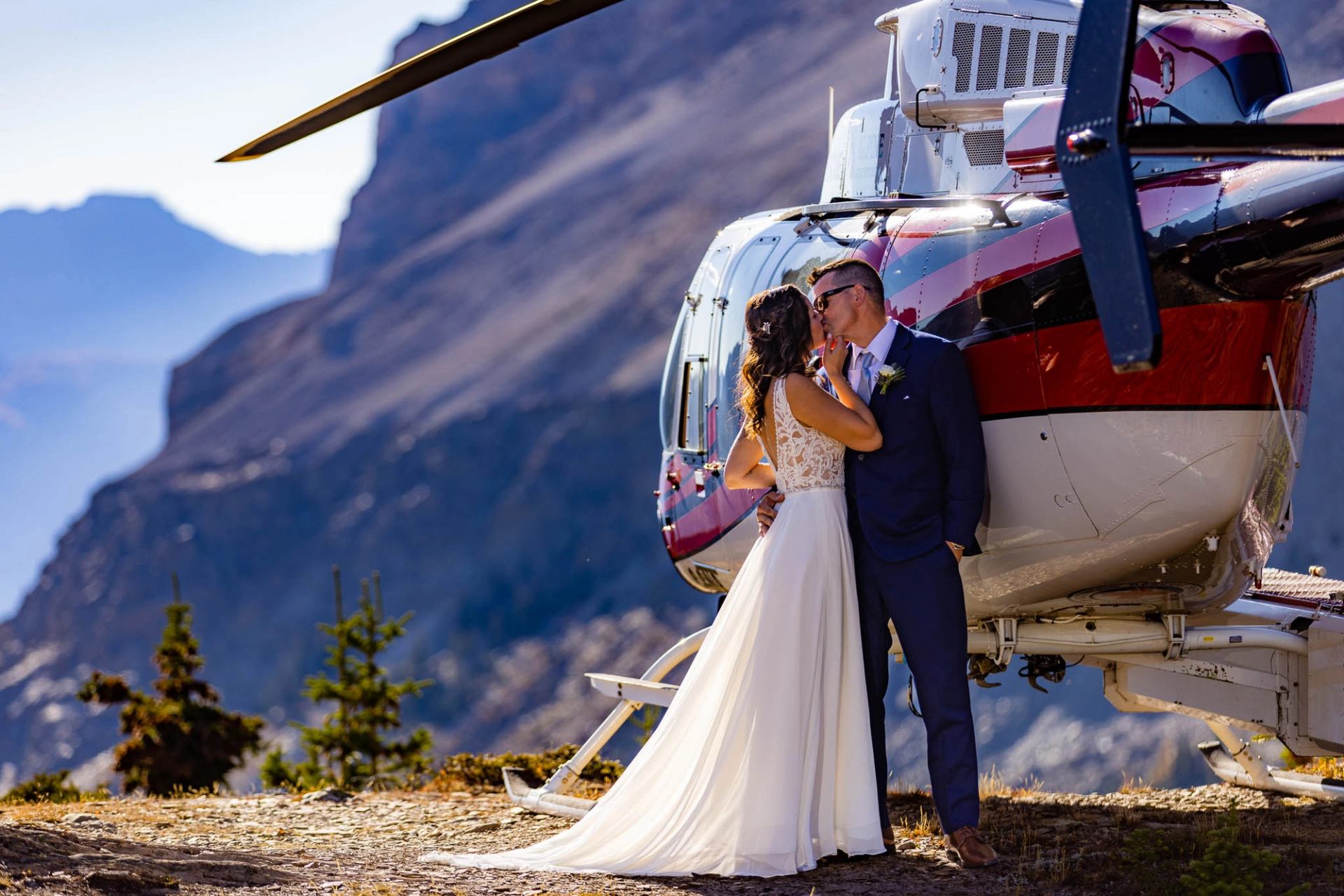 Couple in wedding attire standing beside a helicopter on a mountain landing spot.