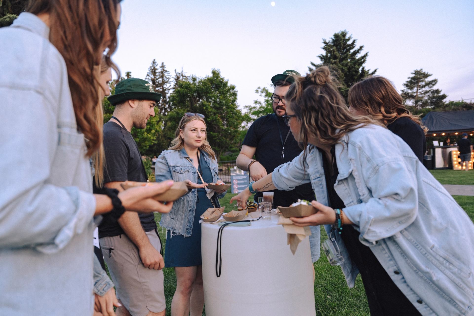 Group of attendees share food and drinks around a standing table outdoors.
