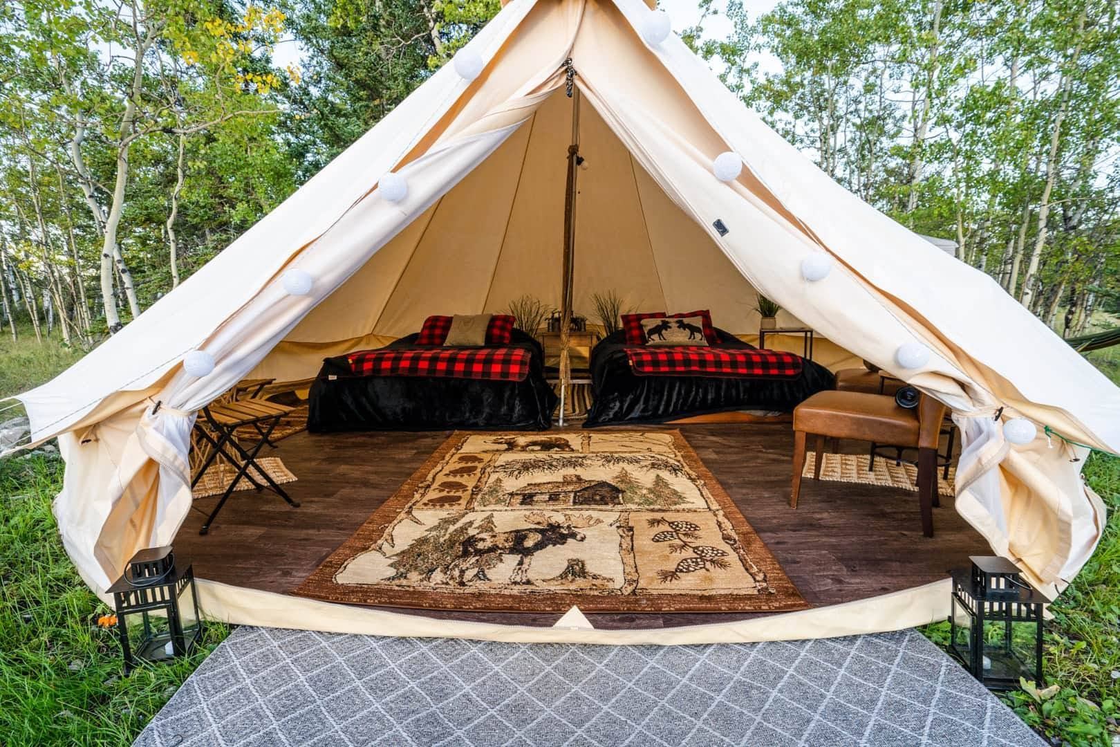 Canvas glamping tent with two plaid-covered beds, rustic rug and forest backdrop.