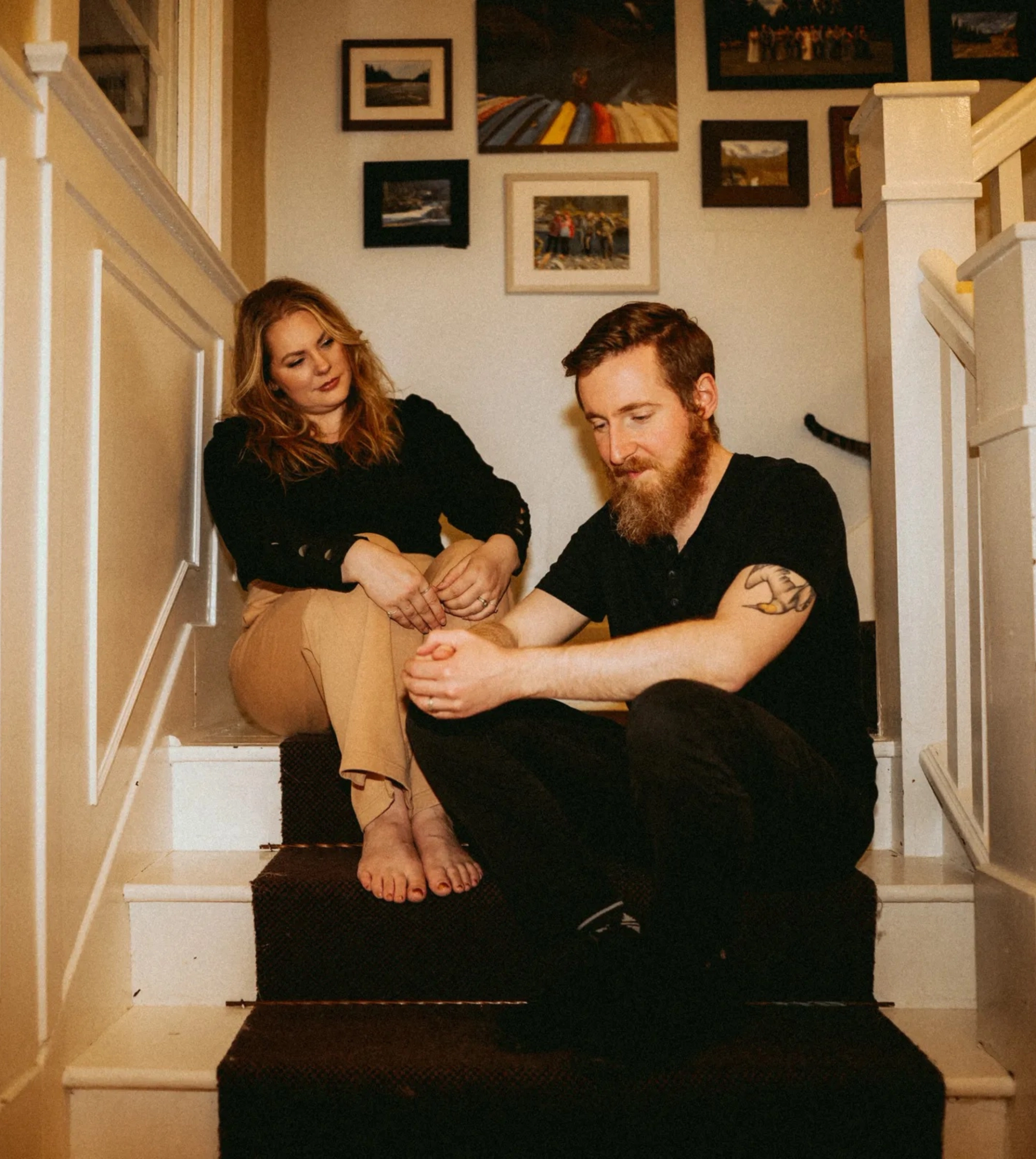 Two adults sit on carpeted stairs in a hallway with framed photos on walls.