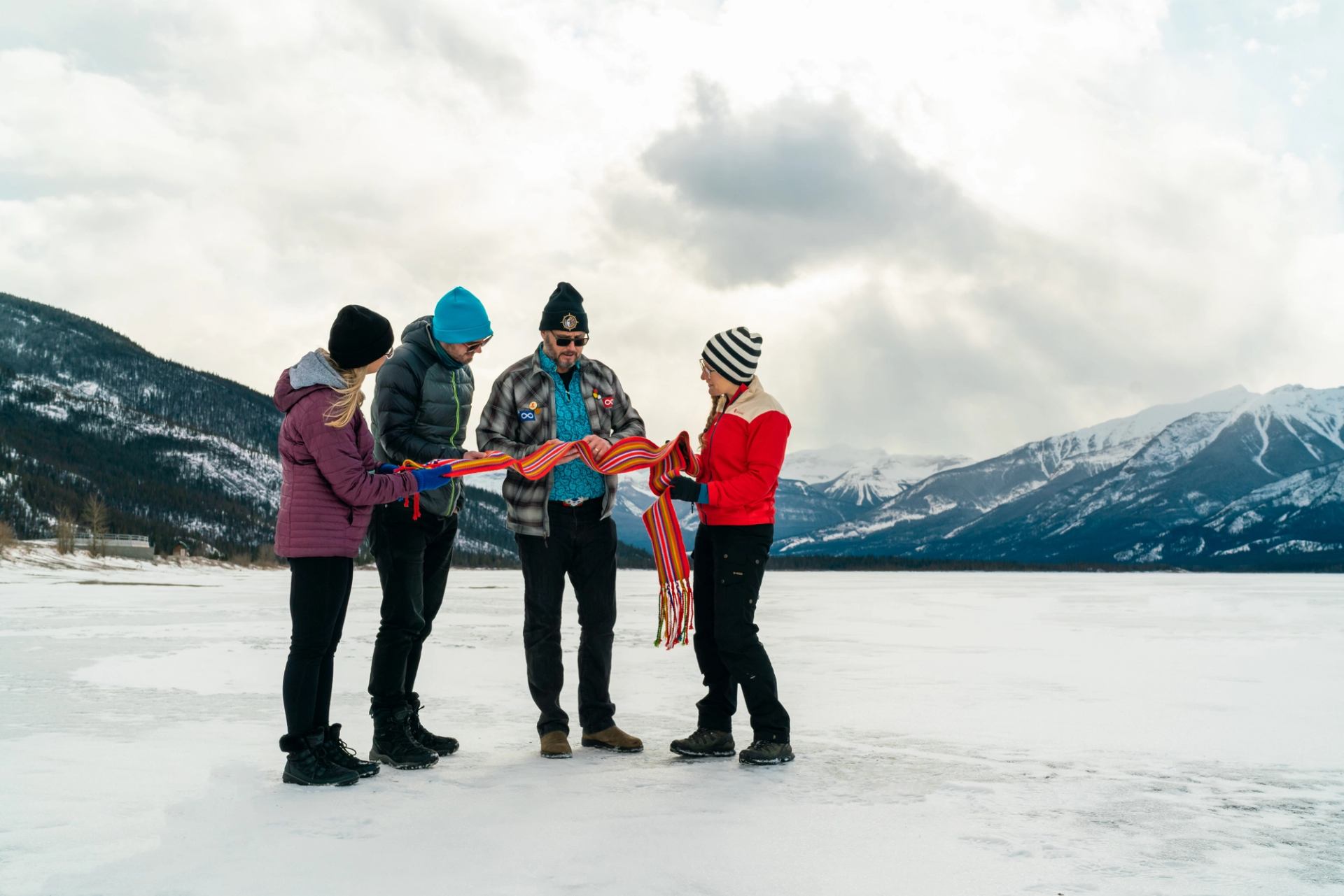 Four people in winter gear hold colorful cloth strips on snow with mountains and cloudy sky behind