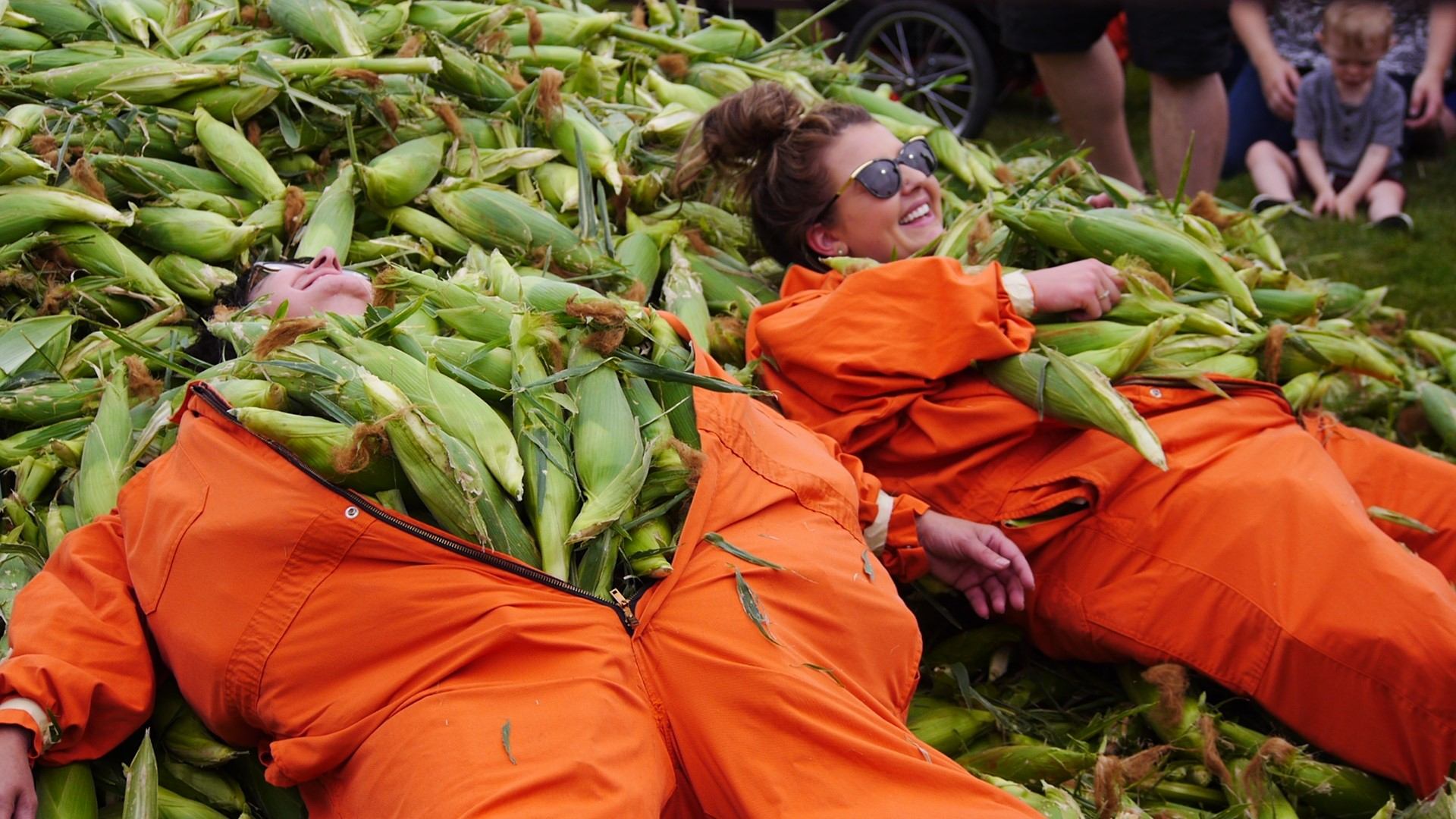 People in orange coveralls lying in a pile of corn cobs.