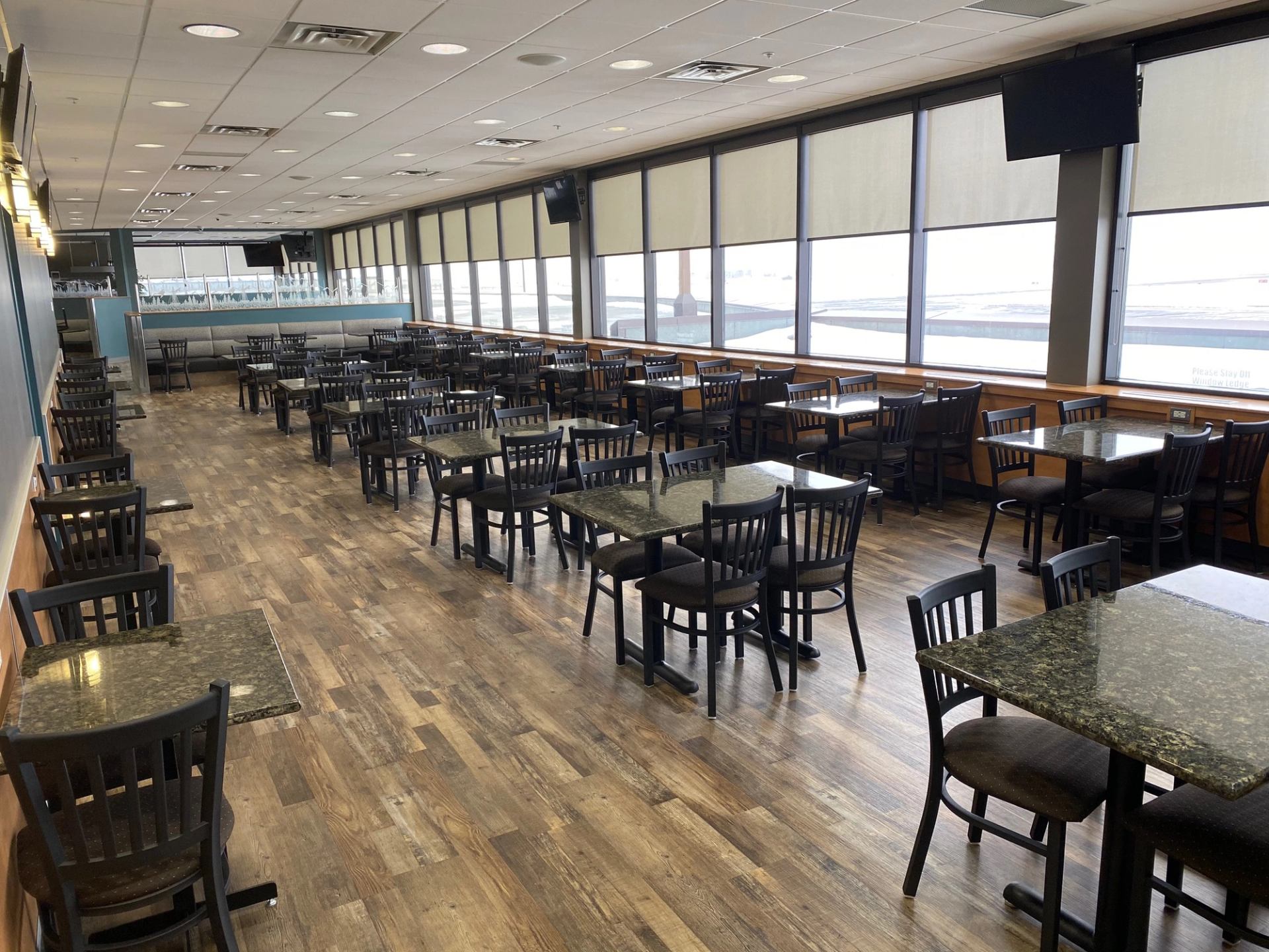 Empty restaurant dining area with rows of dark tables, chairs and large window blinds.