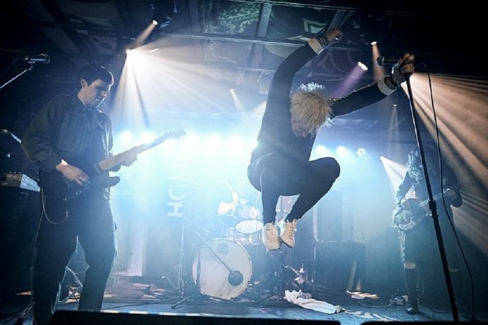 Rock band performs onstage as vocalist jumps midair under bright stage lights.