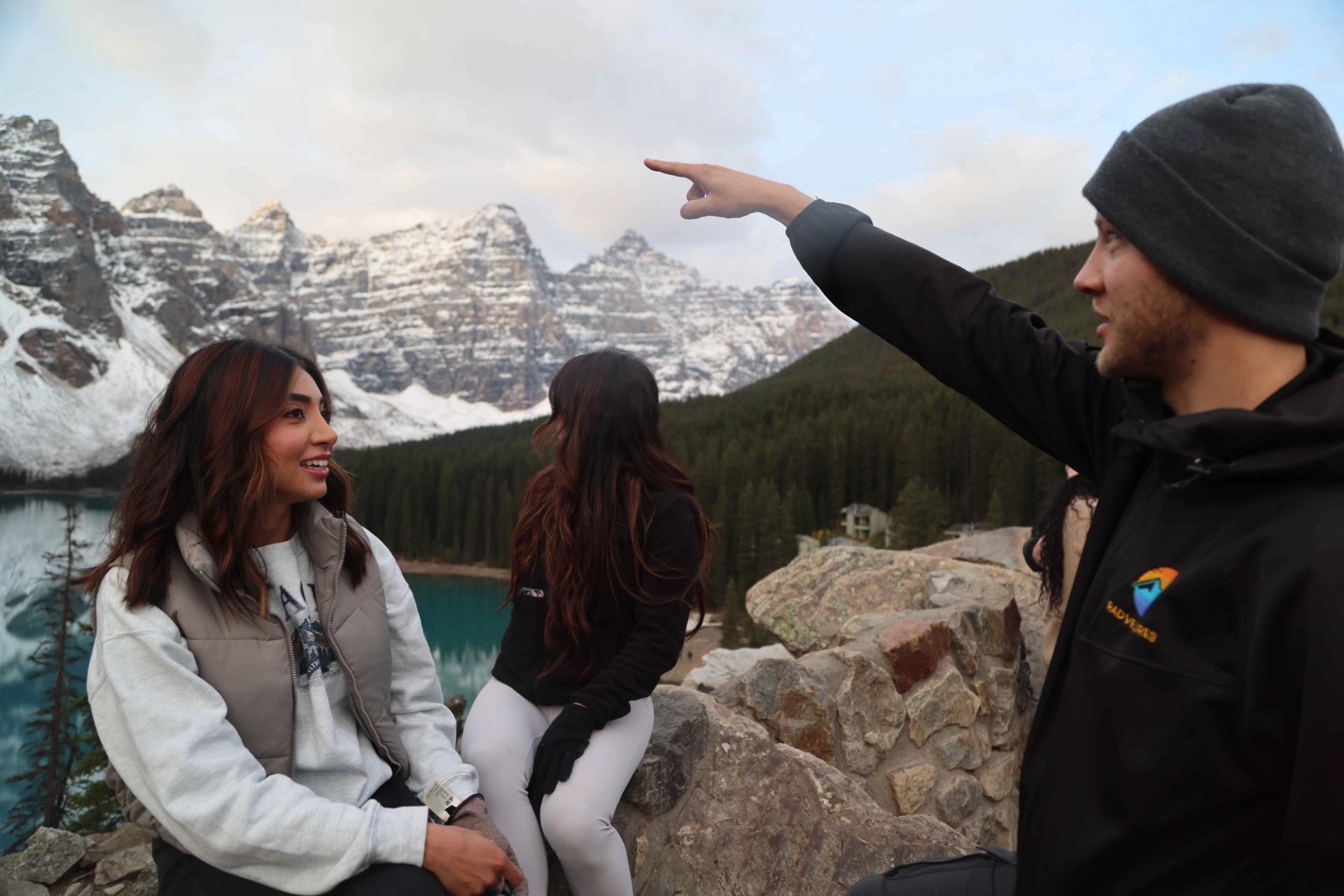A man points toward majestic snow-capped mountains and a turquoise lake while two women look on.