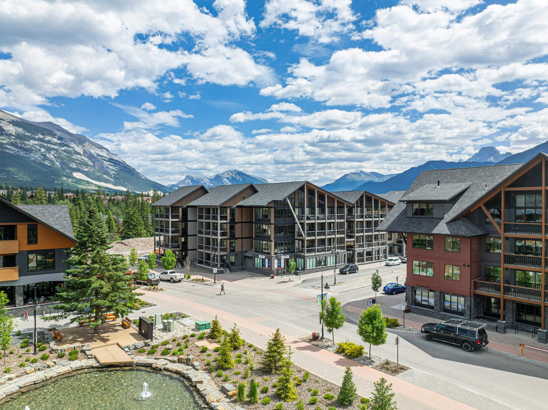 Mountain town buildings surround a street beside a landscaped pond under a cloudy sky.