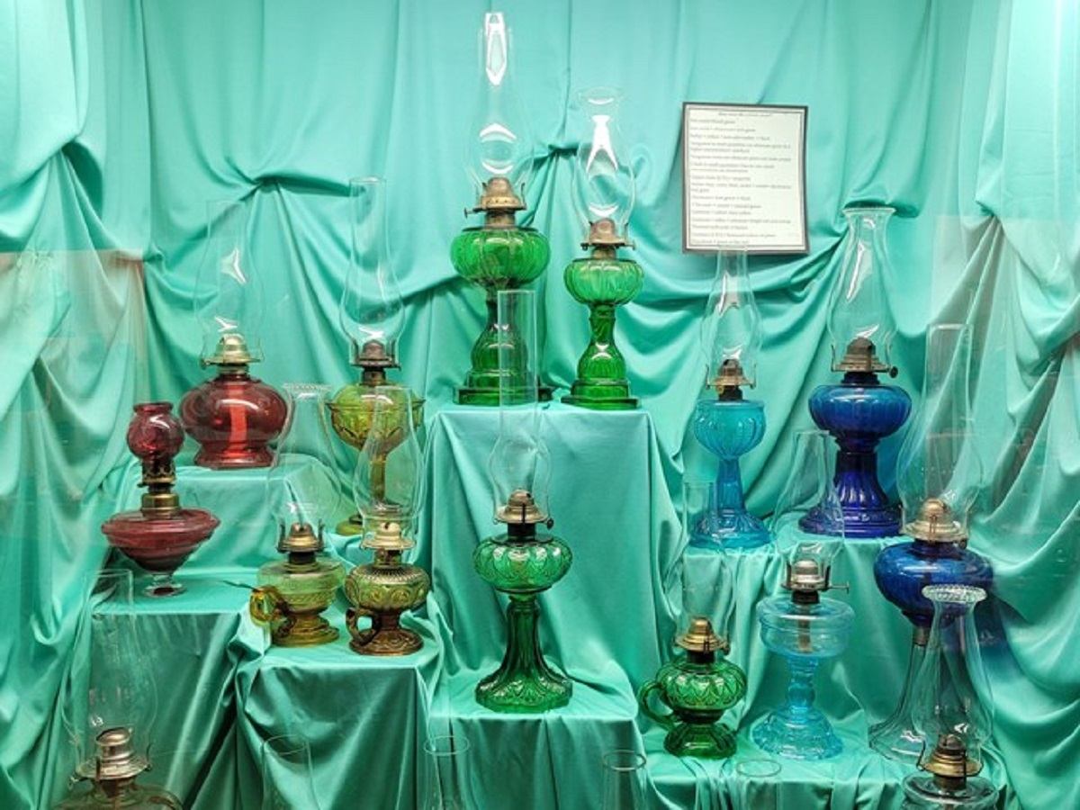 A display of colorful vintage oil lamps in green, blue, red, and other shades, arranged on a teal fabric backdrop.