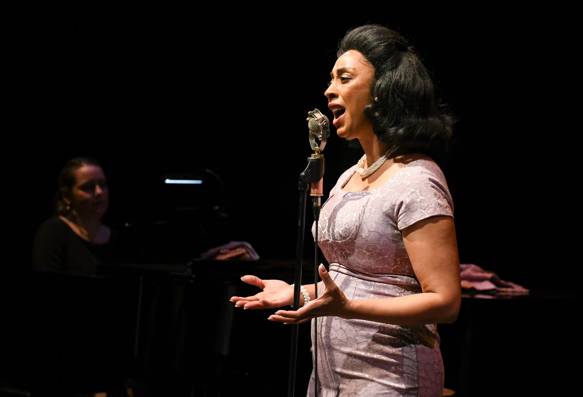Singer in vintage-style dress performs at microphone, pianist playing in dimly lit background.