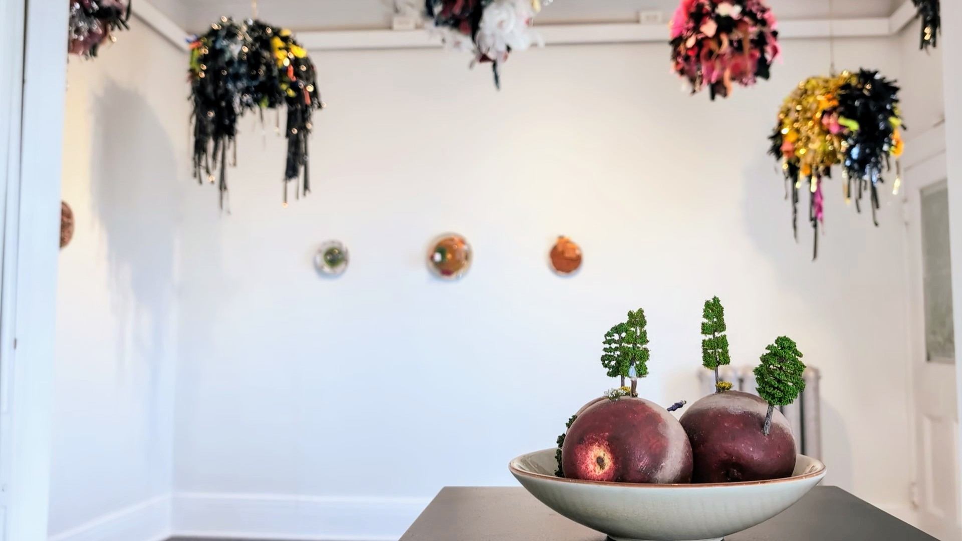 Pictured is a bowl of apples with tiny green trees and miniature people walking on them. In the background there are large scale pompoms 