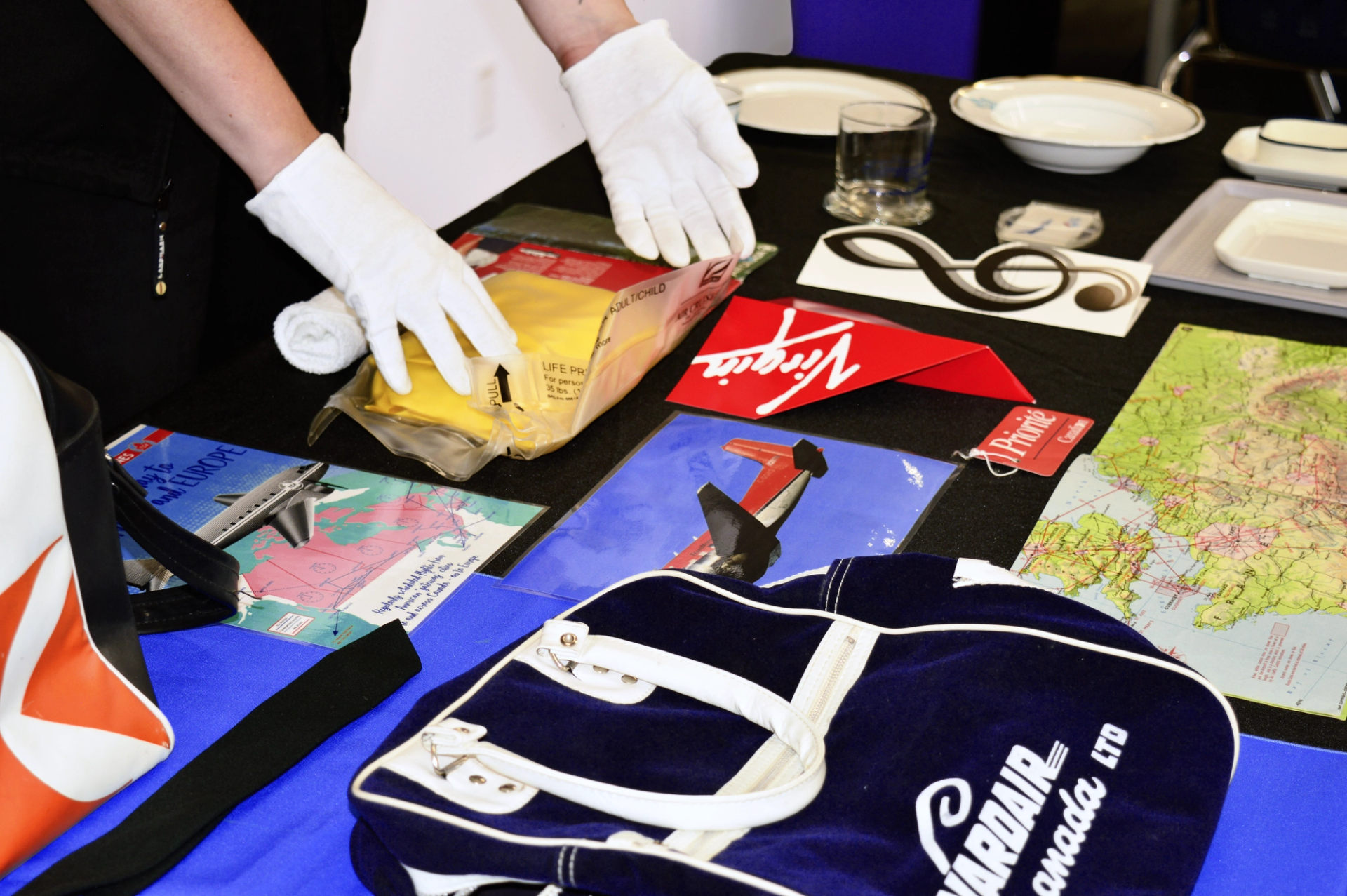Gloved hands arranging a collection of airline memorabilia, including a Virgin logo, a Wardair Canada bag, and a life vest.