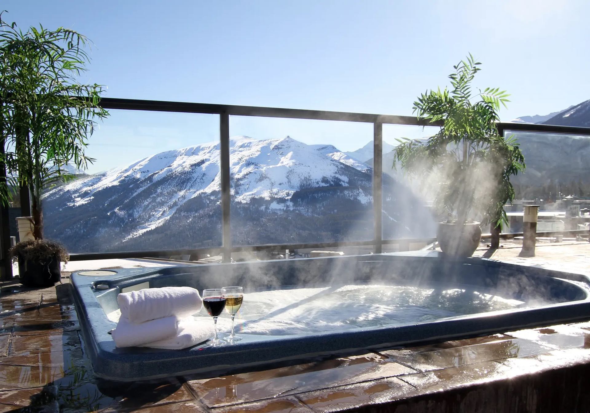 Steaming hot tub on patio with wine, towels, and snowy mountain view.
