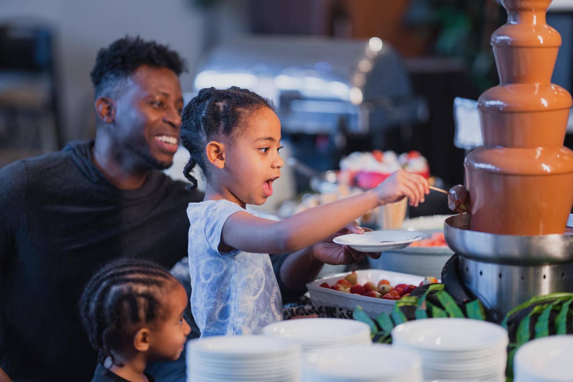 An excited young girl dips fruit into a chocolate fountain, with a smiling man and another child nearby.