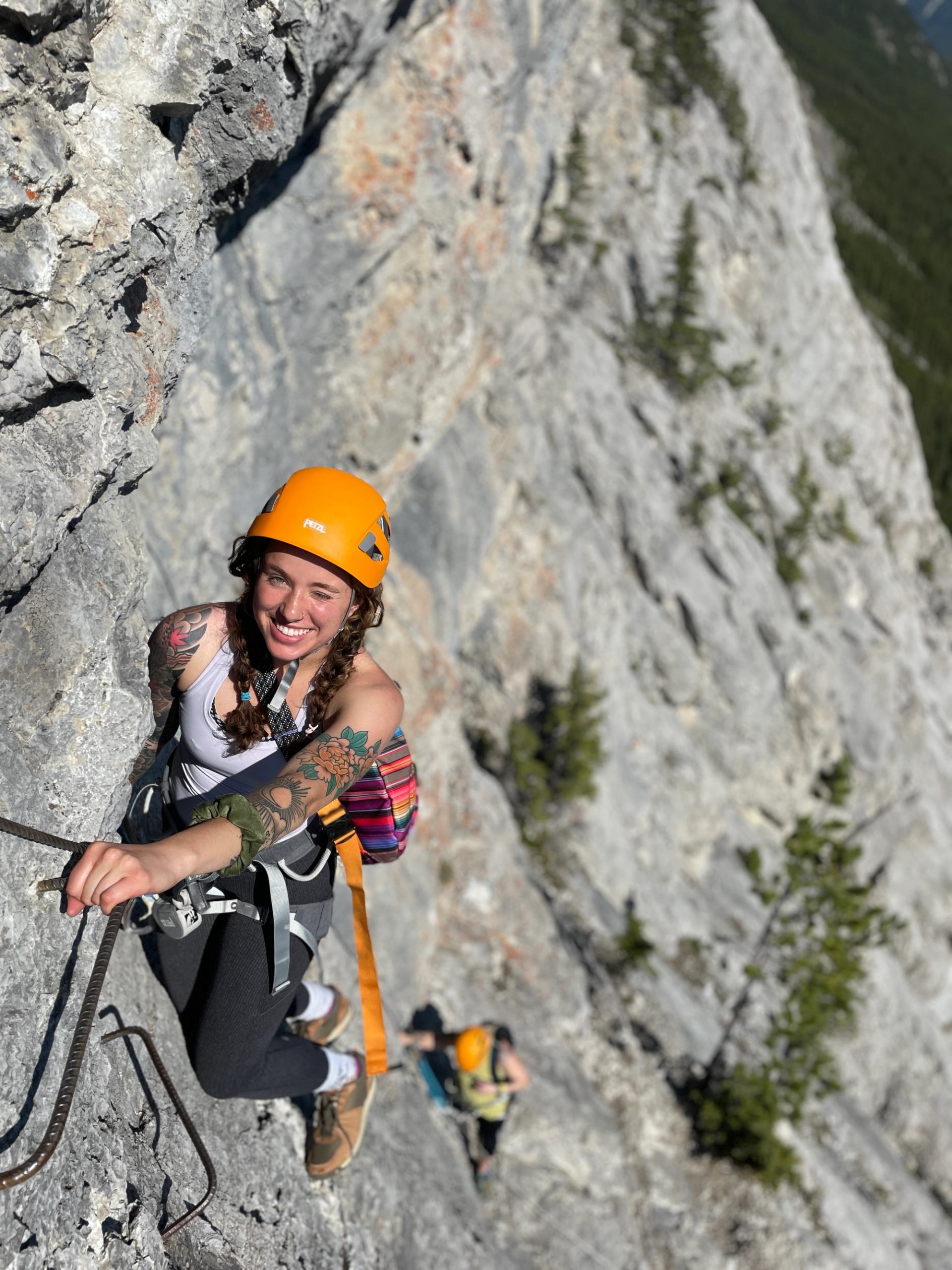 A smiling woman in an orange helmet climbs a via ferrata on a rocky mountain face, with another climber below.