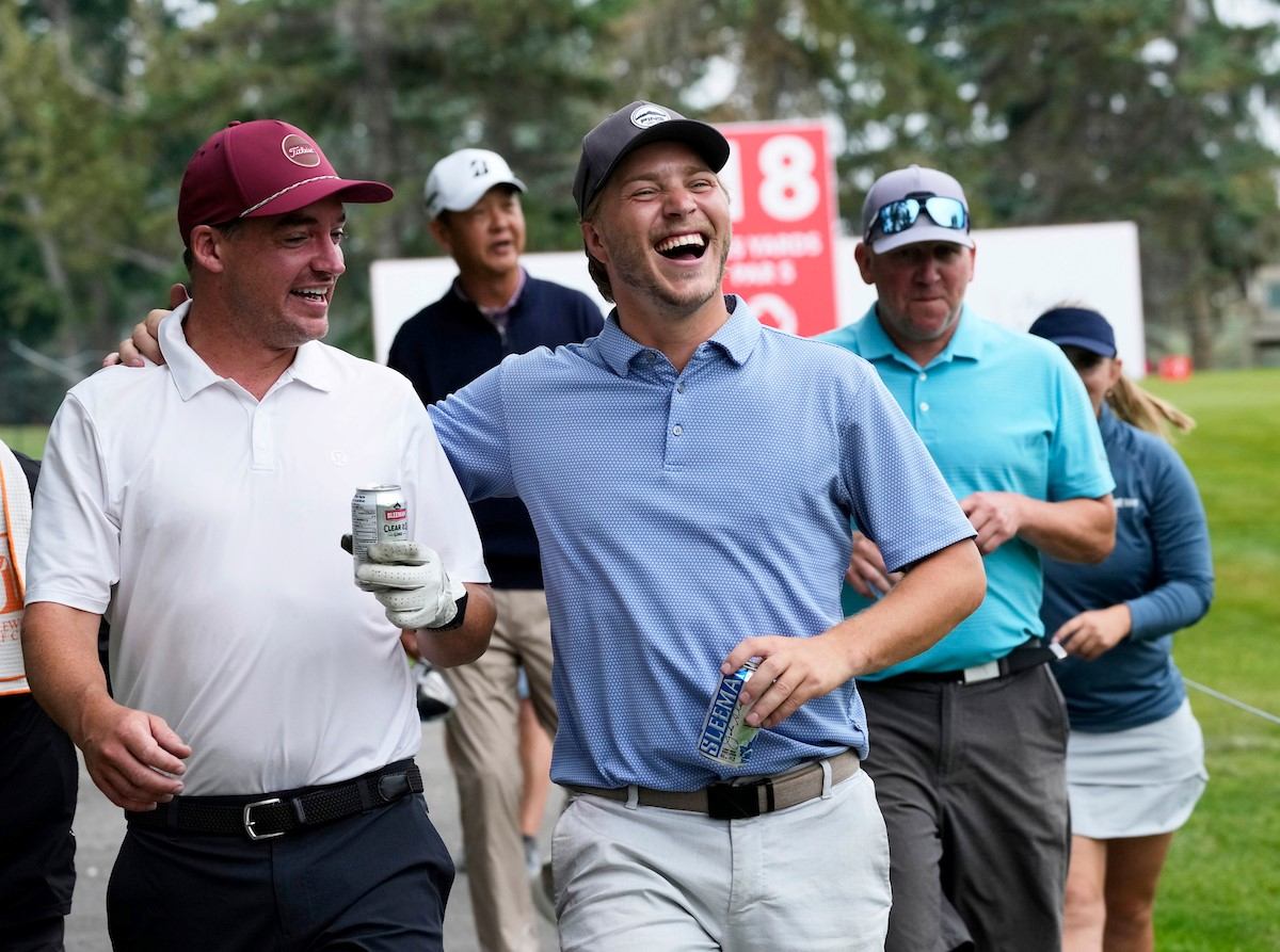 Golfers celebrating on a course, one holds a beverage can and wears a cap.