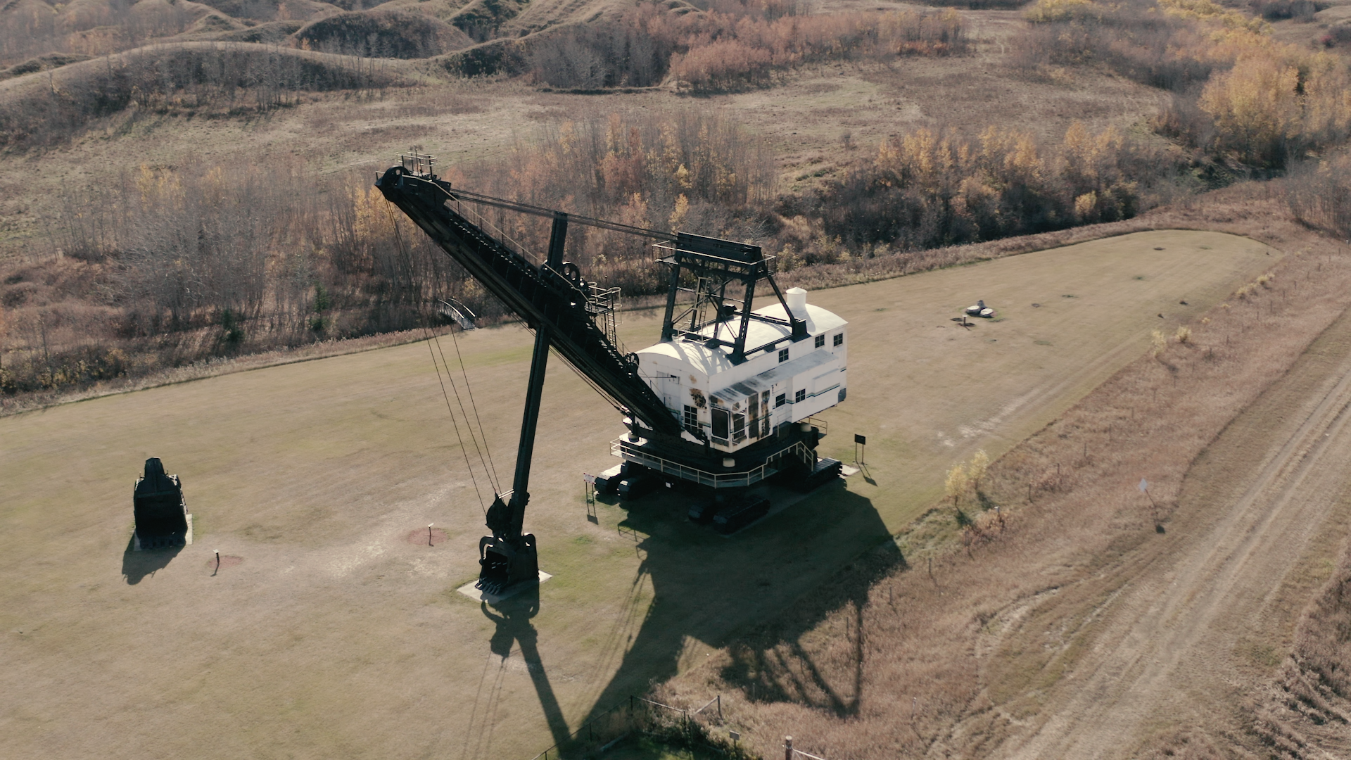 Large historic mining dragline sits on open grassy field beside dirt road and trees.