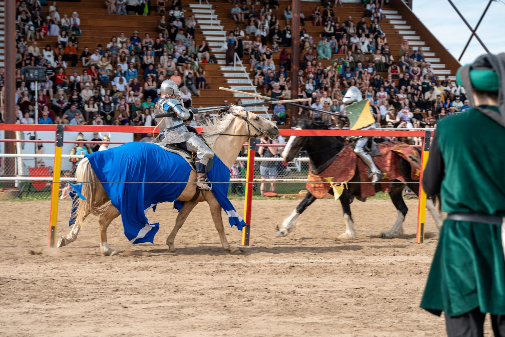 Armored fighters on horseback competing in a medieval jousting match inside an arena with spectators.
