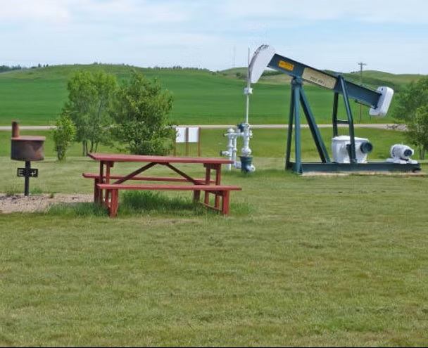 A picnic table and barbecue stand on a grassy campsite with rolling hills and open countryside.