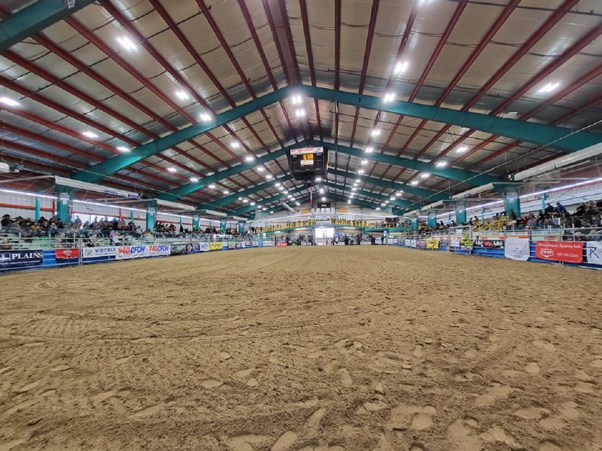 Indoor arena with a dirt floor, spectators in the stands, and a scoreboard.