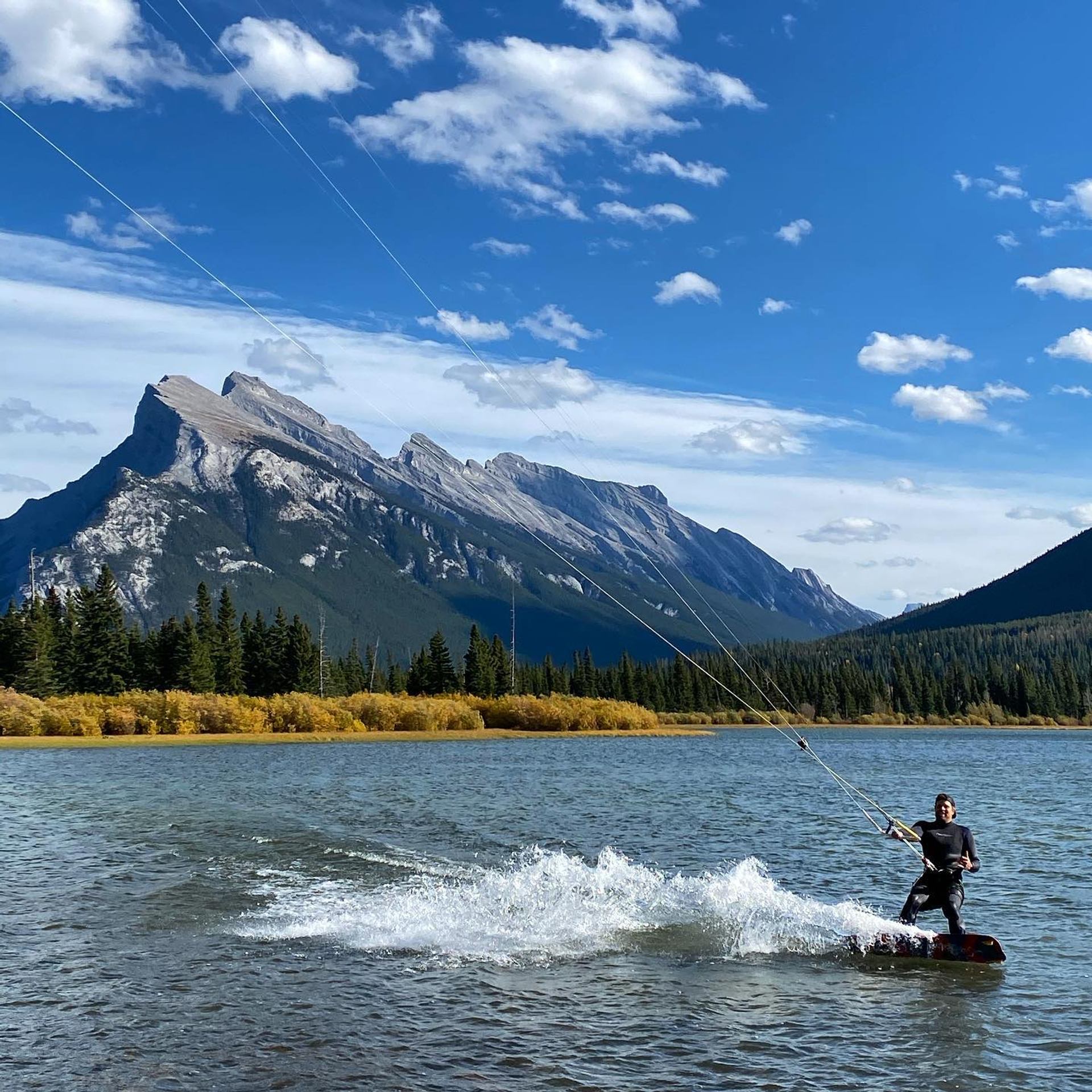 Kiteboarder skims across a mountain lake, spraying water with rugged peaks and blue sky in the background.