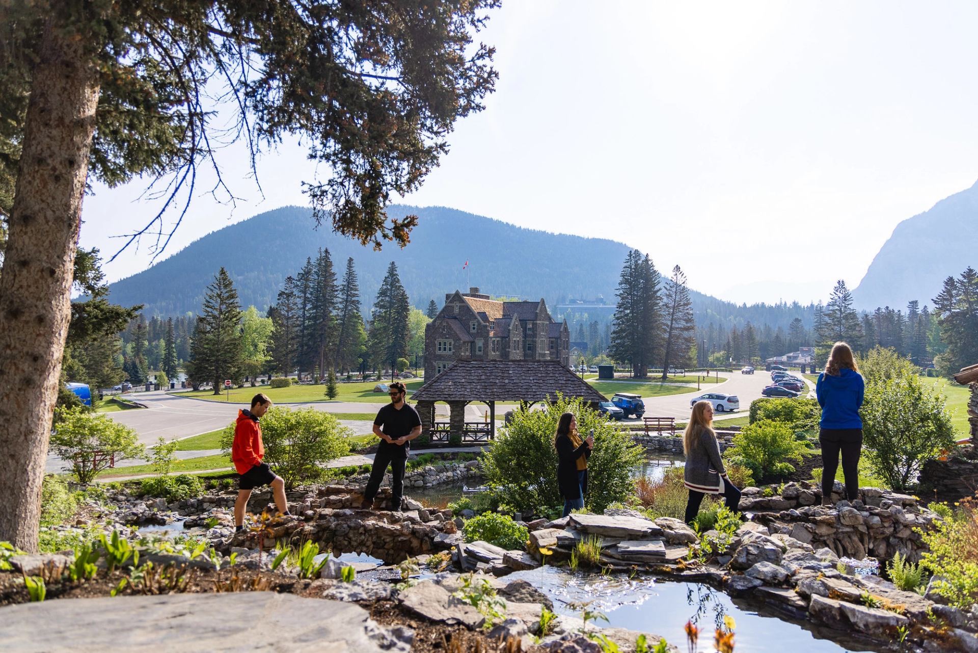 People stand on rocks near a pond with mountains, trees and stone building behind.