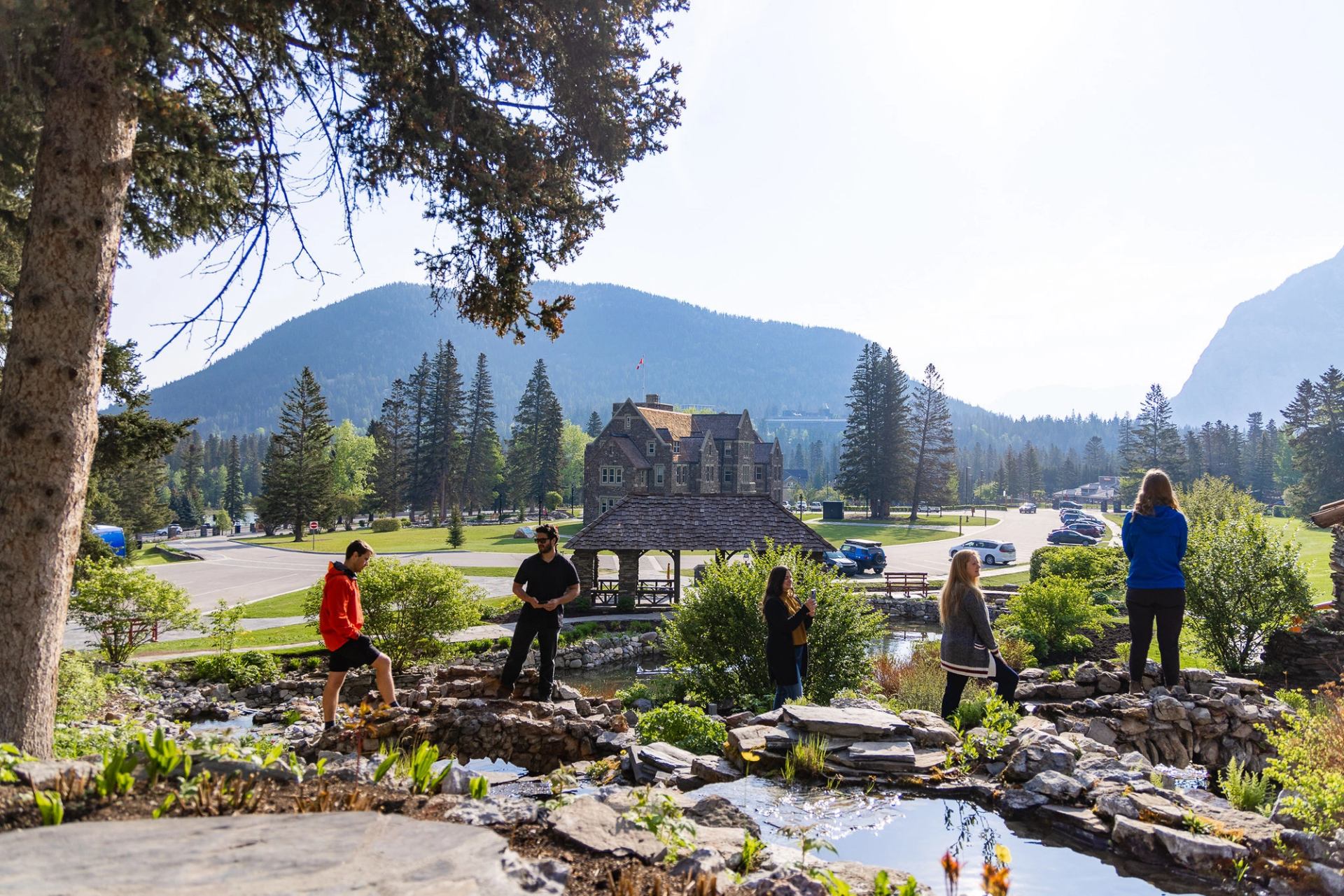 People stand on rocks near a pond with mountains, trees and stone building behind.