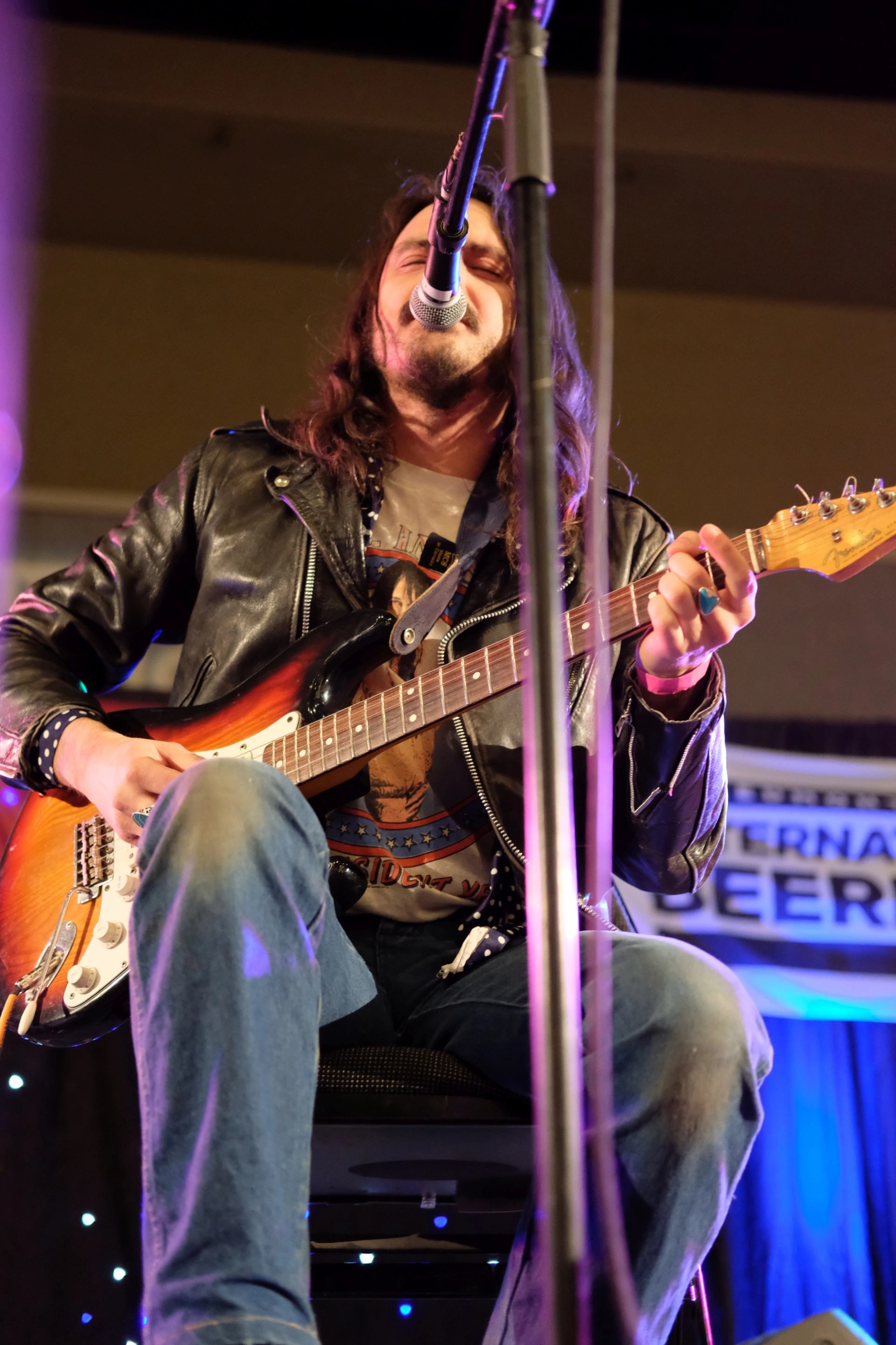 Guitarist in leather jacket sits onstage, playing electric guitar and singing into microphone.