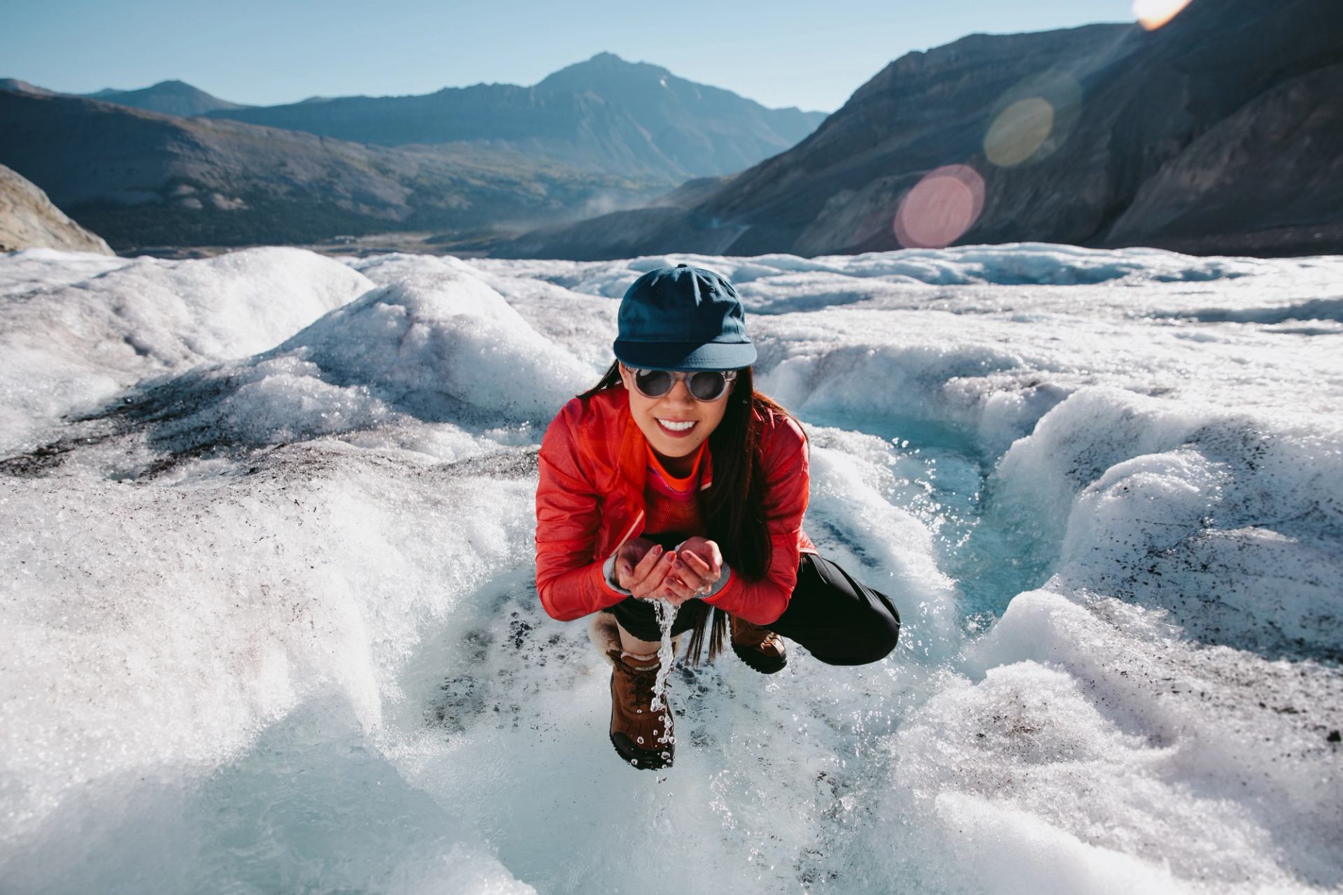 Person in red jacket crouches on glacier, holding melting ice over blue water.