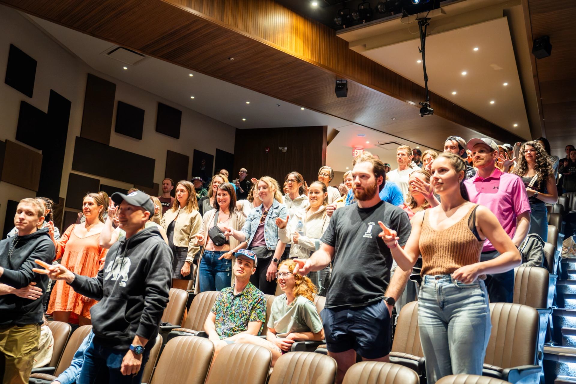 Audience members stand in a theatre, gesturing with their hands during an event.