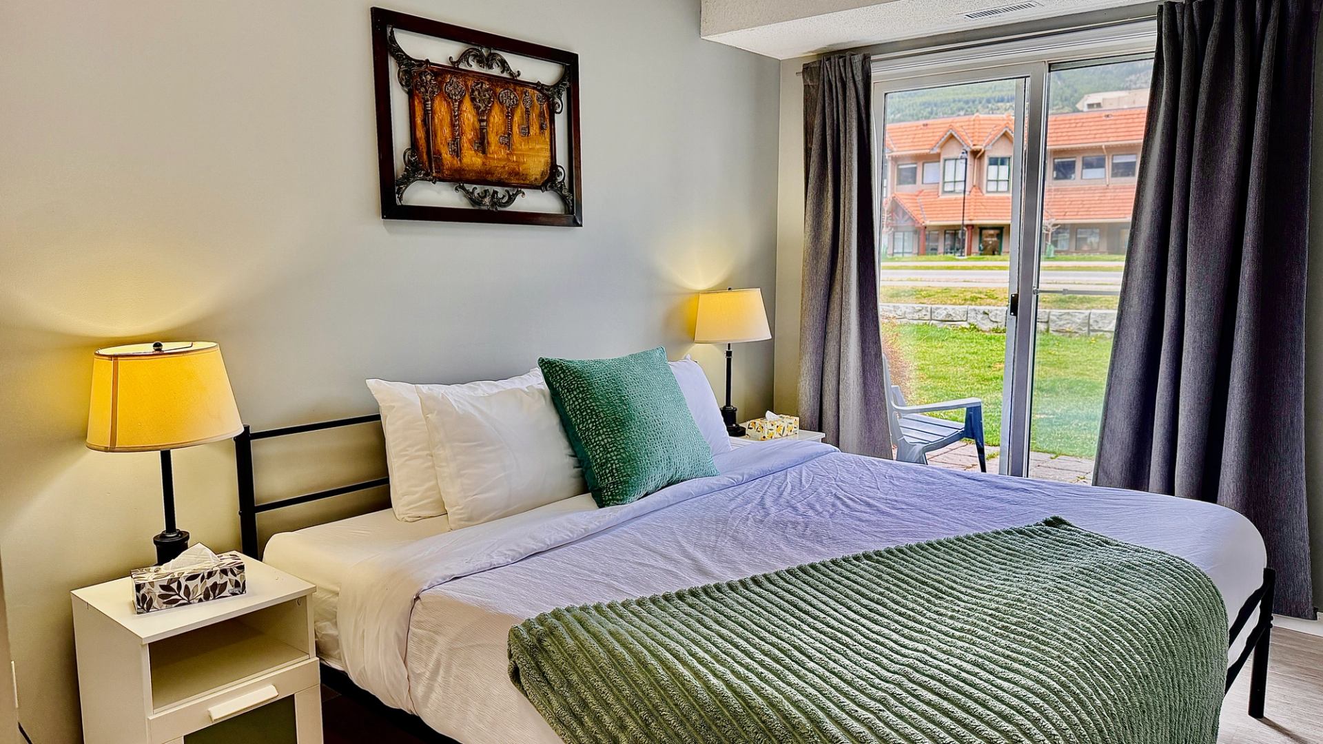 Bedroom with green bedding, bedside lamps and glass doors overlooking a lawn.