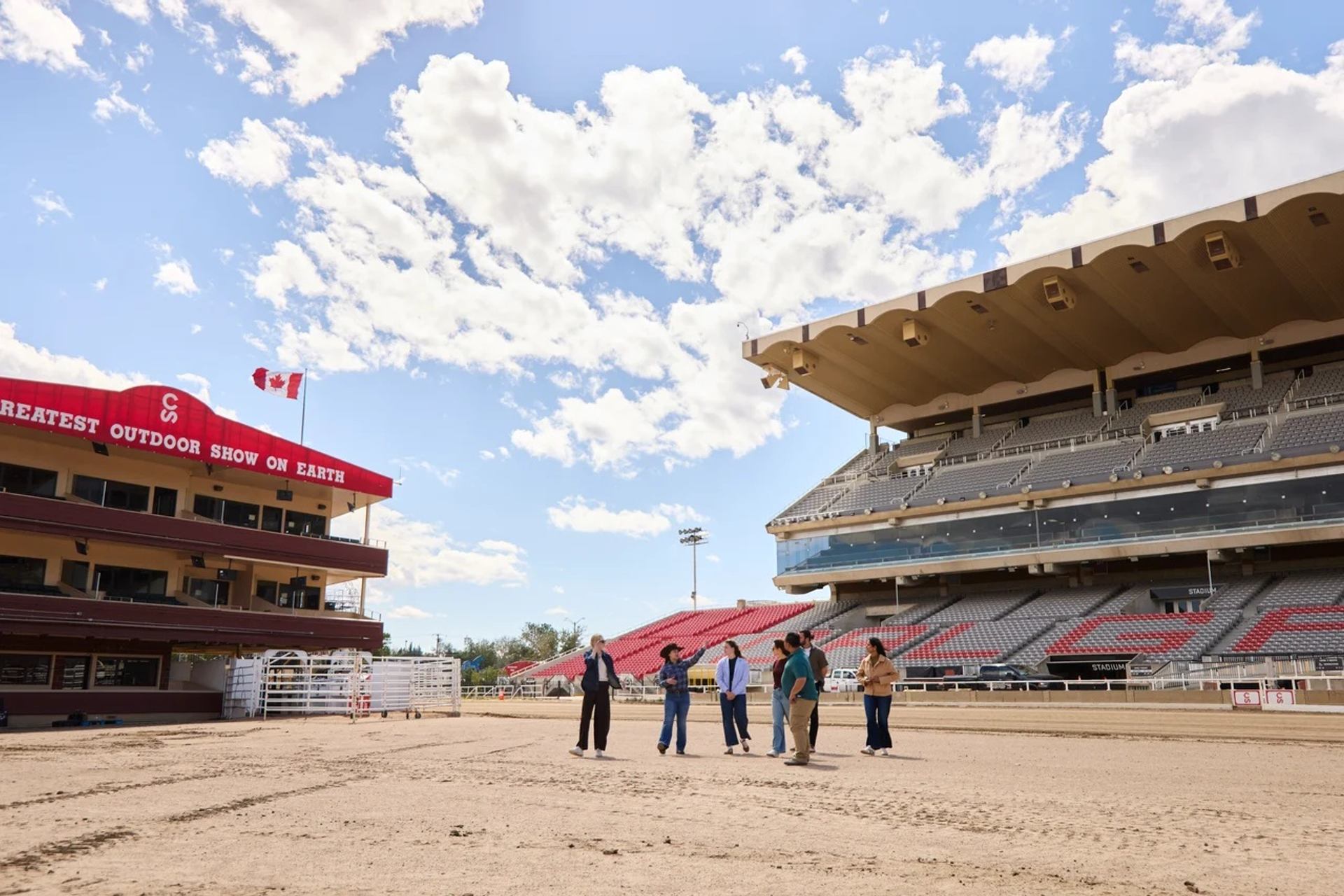 Group of people stand on rodeo arena dirt between grandstands under partly cloudy sky.